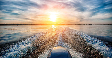 A view from the back of a boat with the motor creating waves over the water as the sun sets over the land in the distance.