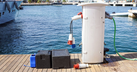 Two black marine batteries are sitting next to a power source on the dock of a marina. The water beyond the dock is dark blue.