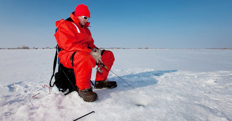 A man wearing insulated red clothing and sunglasses sitting on a chair with a pole while ice fishing during the day.