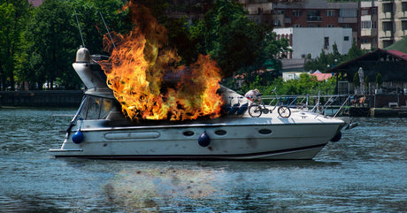 A yacht sitting on the water that’s caught fire and started to char black with the shore in the background.