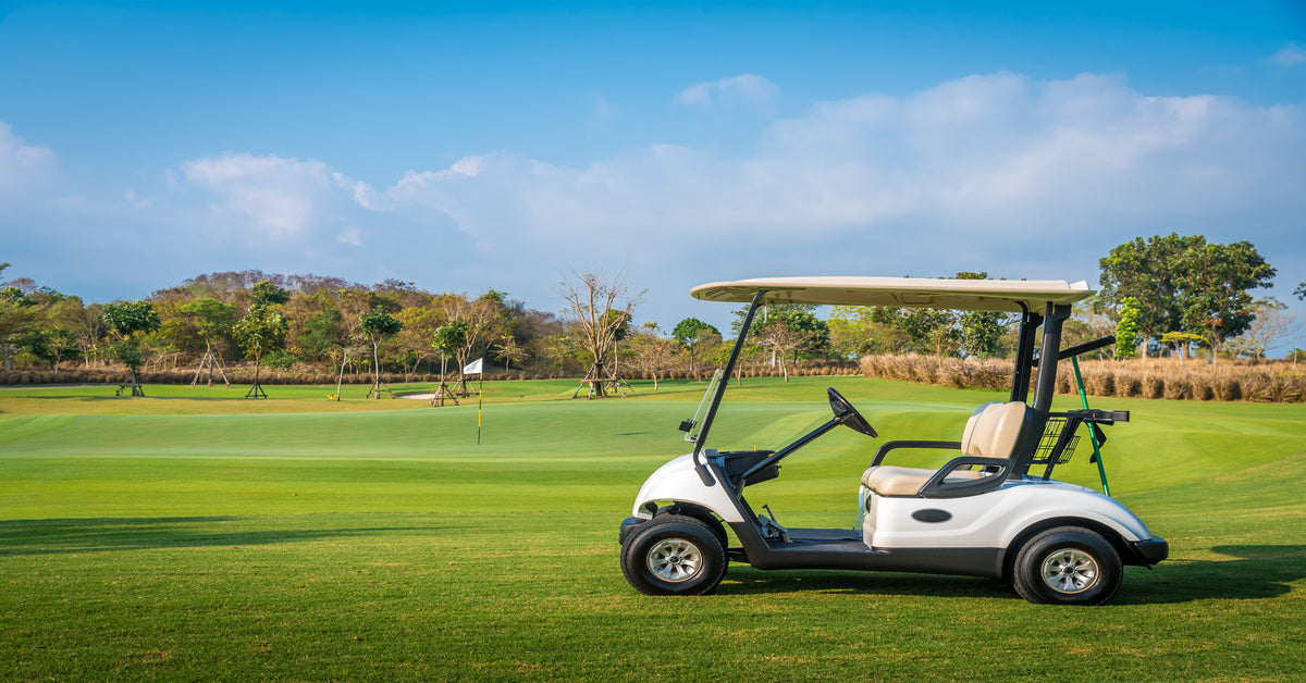 A golf cart parked on a green in front of a flag. The sky above is blue and cloudy and there are trees in the distance.
