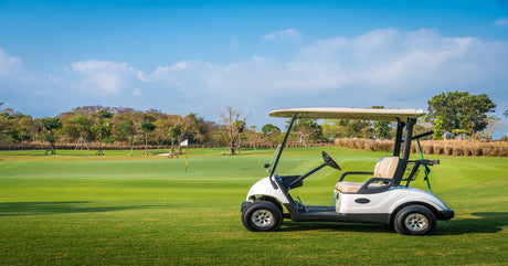 A golf cart parked on a green in front of a flag. The sky above is blue and cloudy and there are trees in the distance.