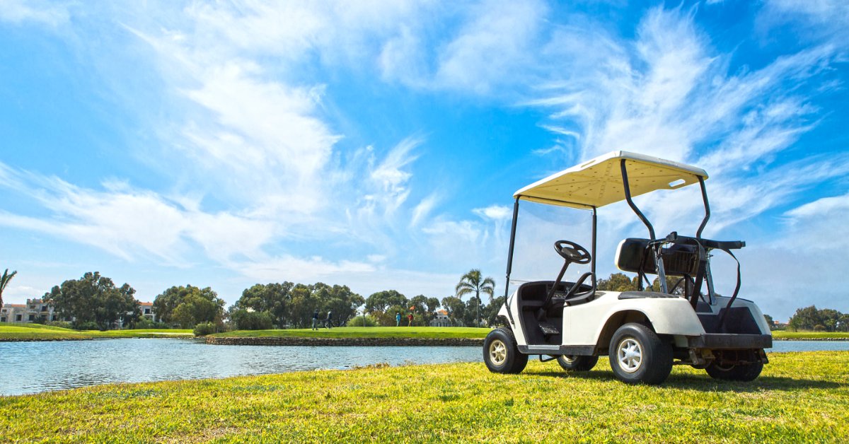 A typical white golf cart sits empty on a grassy area next to a pond. The blue sky above contains some clouds.
