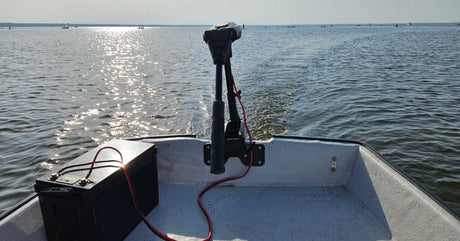 A black trolling motor is attached to a white boat that's on a large body of water. A battery uses a red cord to power the motor.