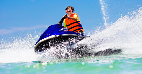A teenage boy wearing a bright orange life vest and sunglasses while driving a blue jet ski on water.