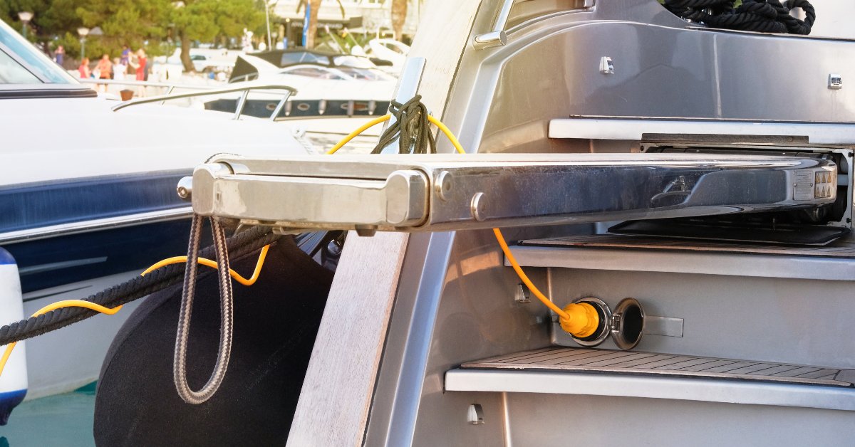 A yellow charging cable is plugged into a port that's on the step of a boat. The boat sits at a marina with other boats.