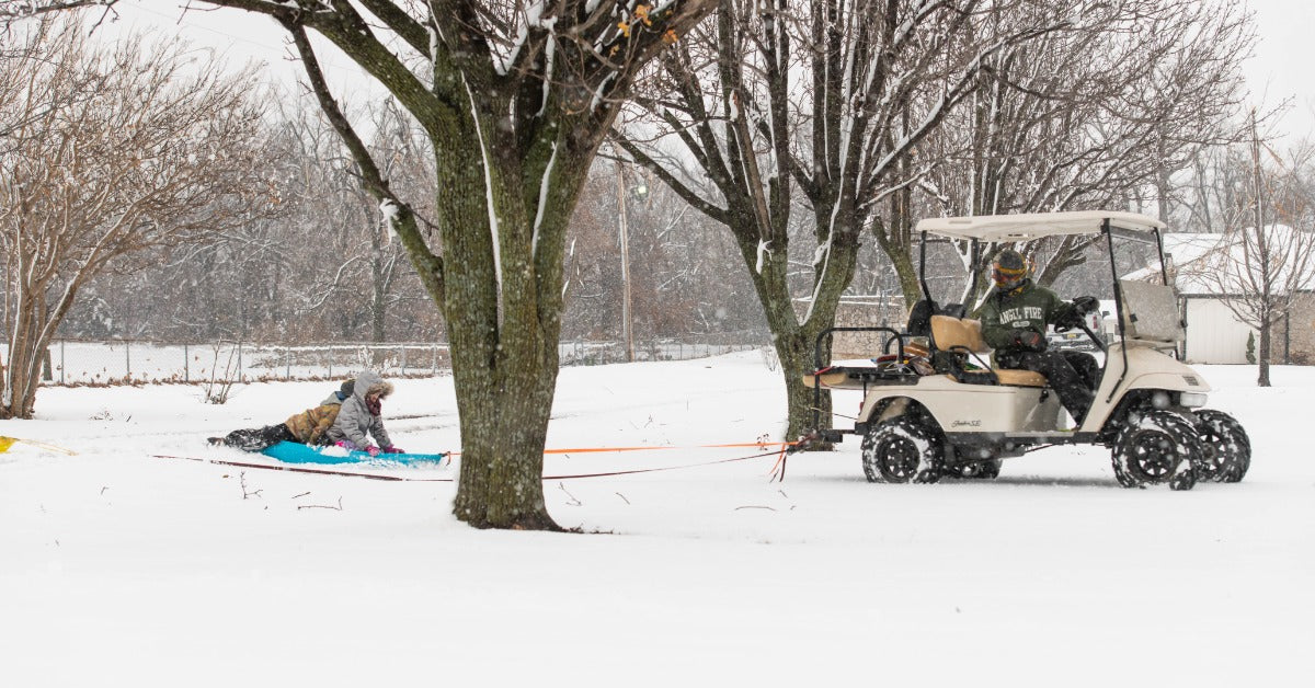 A man is driving a white golf cart across snow-covered ground and pulling a sled with two kids on it.
