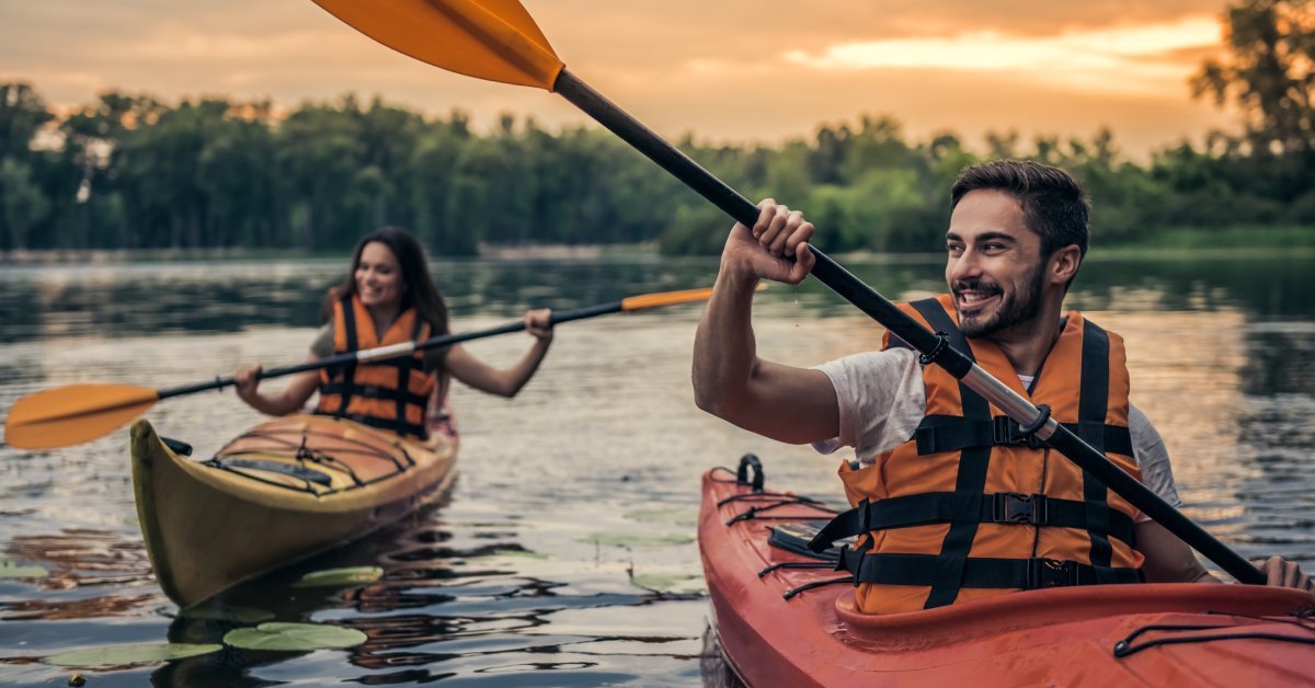 A man and a woman wearing life jackets while kayaking on the water at sunset. Both are smiling.