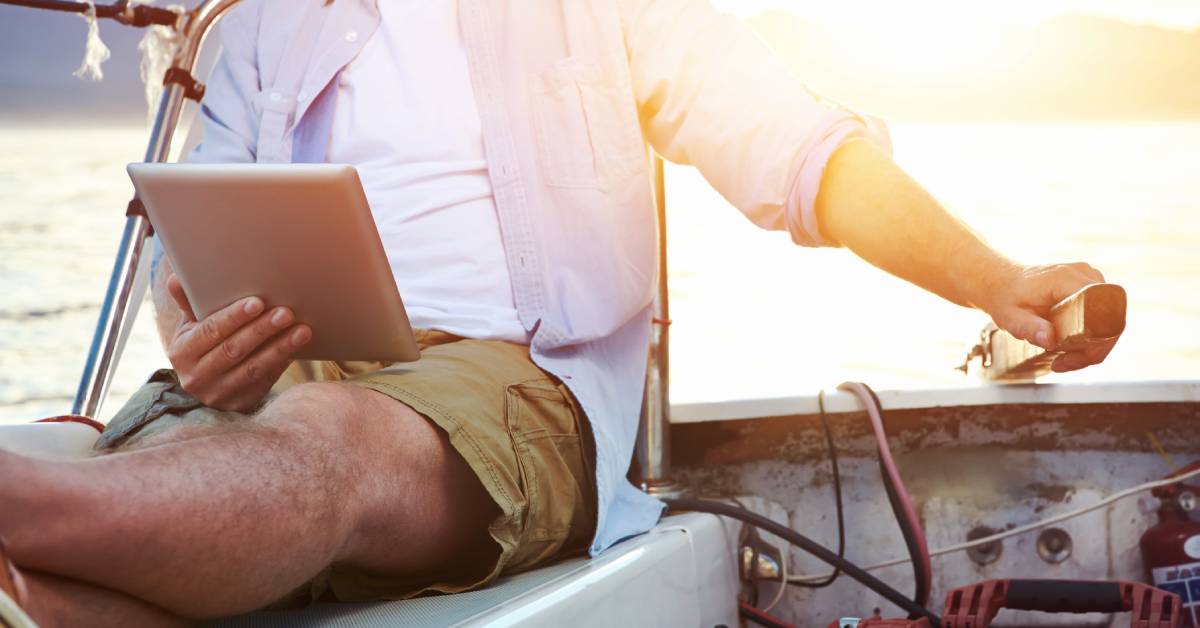 A man wearing khaki shorts and a pink shirt sitting on a boat with a computer tablet with his hand on the rudder.