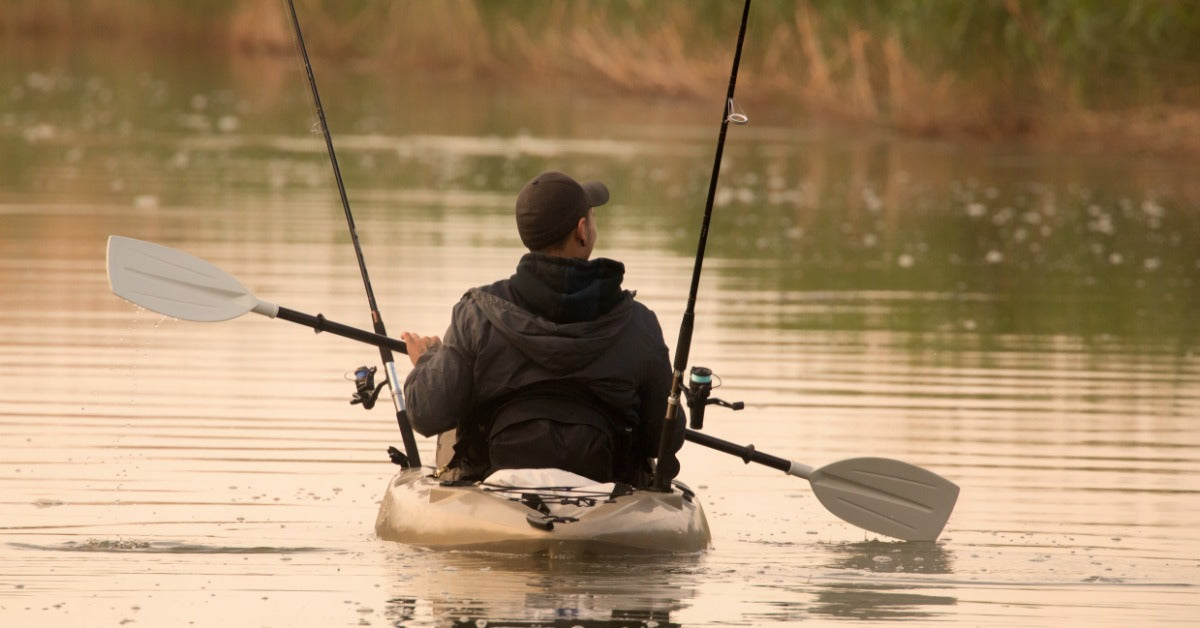 A man wearing a black jacket and baseball cap is using a double-sided paddle to move a kayak. Fishing poles are in the kayak.