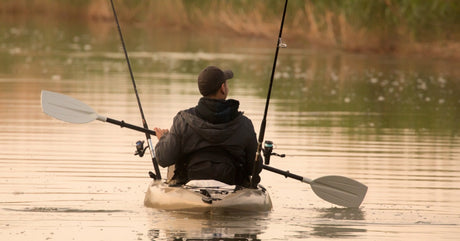 A man wearing a black jacket and baseball cap is using a double-sided paddle to move a kayak. Fishing poles are in the kayak.