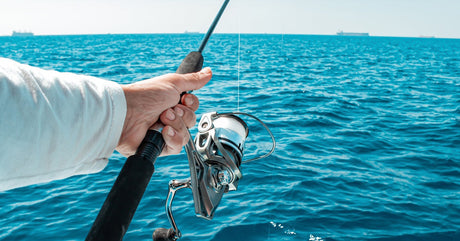 A person holding out a long black fishing rod by its handle, with the reel and line free above the blue ocean.