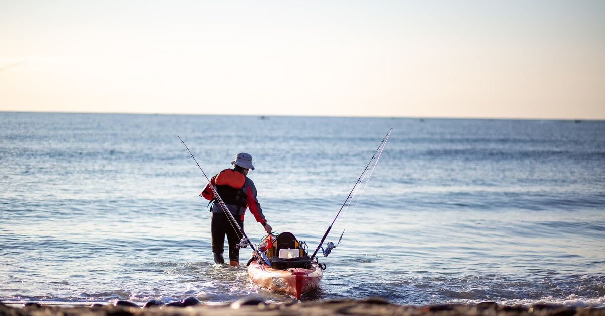 A person with intermediate fishing gear pulling their orange kayak out onto the water from a rocky beach.