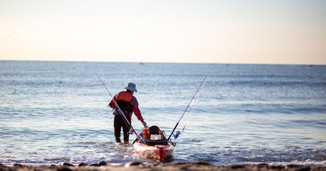 A person with intermediate fishing gear pulling their orange kayak out onto the water from a rocky beach.