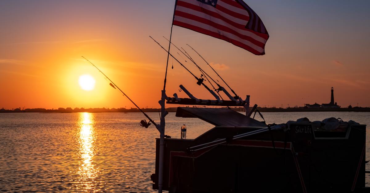 A boat with many rods hanging over its side sitting on the water at sunset. A pole with a US flag hangs over the boat.
