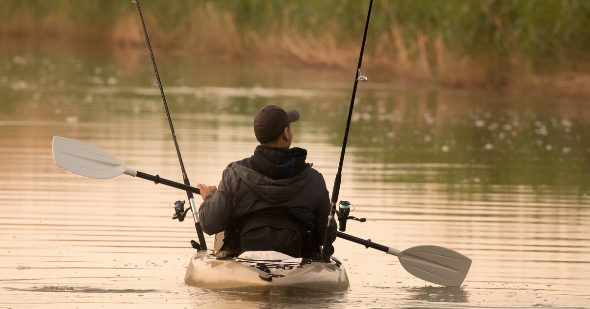 An angler rowing on a tan kayak across calm waters. Two rods hang from either side of the fisherman's kayak.
