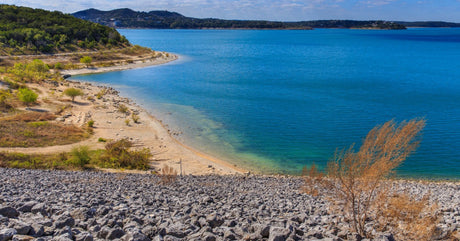Canyon Lake in the Texas hill country on a clear day. A rocky hill leads down to the sandy beach and vivid blue waters.