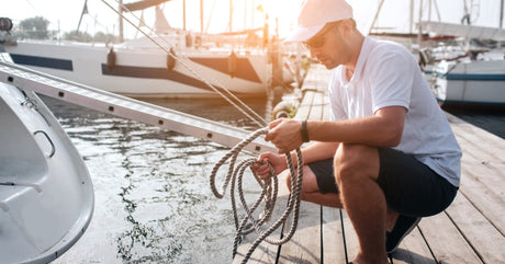 A boat owner kneeling on a dock behind their boat while tidying up a length of simple rope after untying it.