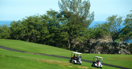 A pair of black-and-white golf carts being driven across a wide golfing green on an island golf course in Hawaiʻi.