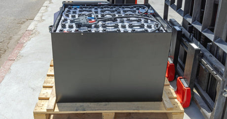 A large lead-acid battery with a charcoal gray case and many covered terminals sitting on top of a wooden pallet.