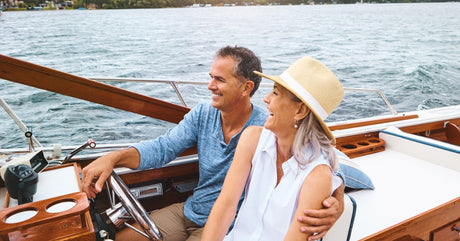 A couple sitting at the wheel of a luxury boat with wooden paneling. They are both smiling and having fun.